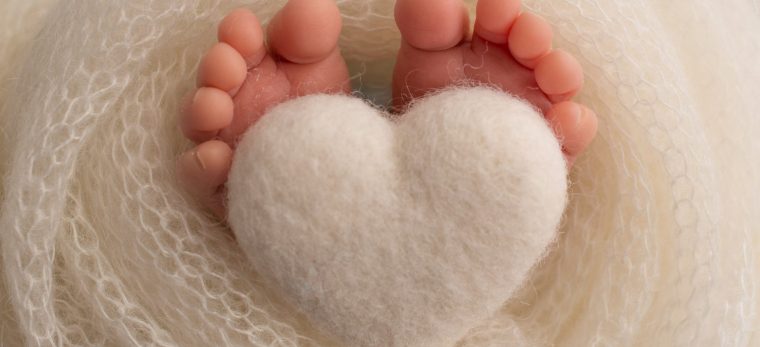 The feet of a newborn baby are wrapped in a knitted blanket. The fingers of a newborn baby are holding a white knitted heart. High quality photo