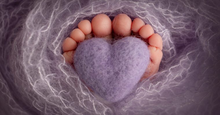 The tiny foot of a newborn baby. Soft feet of a new born in a purple, lilac wool blanket. Close up of toes, heels and feet of a newborn. Knitted purple, lilac heart in the legs of a baby.